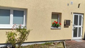 Ein zweistöckiges Gebäude mit sandfarbenen Wänden, einem kleinen Fenster mit roten Blumen und einer Tür, über der die Zahl '2' angebracht ist.