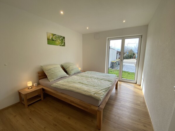 Ein Schlafzimmer mit einem hölzernen Bett, einem Nachttisch, einer Wandleuchte und einer Glastür, die auf einen Balkon führt.