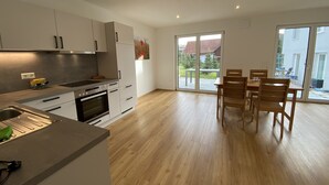 A modern kitchen with white cabinets and a dining area with wooden chairs and a table. - Ferienwohnung in Ravensburg, Neubau, Ruhig, in der Natur, Barrierefrei, Terrasse (Ravensburg)