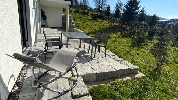 A patio with a table and chairs, a glass balustrade, and a view of a grassy hillside with trees. - Ferienwohnung in Ravensburg, Neubau, Ruhig, in der Natur, Barrierefrei, Terrasse (Ravensburg)