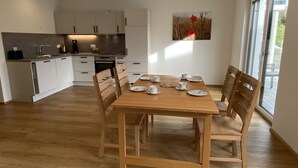A modern kitchen with white cabinets, a wooden dining table set with plates and cups, and a large window providing natural light. - Ferienwohnung in Ravensburg, Neubau, Ruhig, in der Natur, Barrierefrei, Terrasse (Ravensburg)