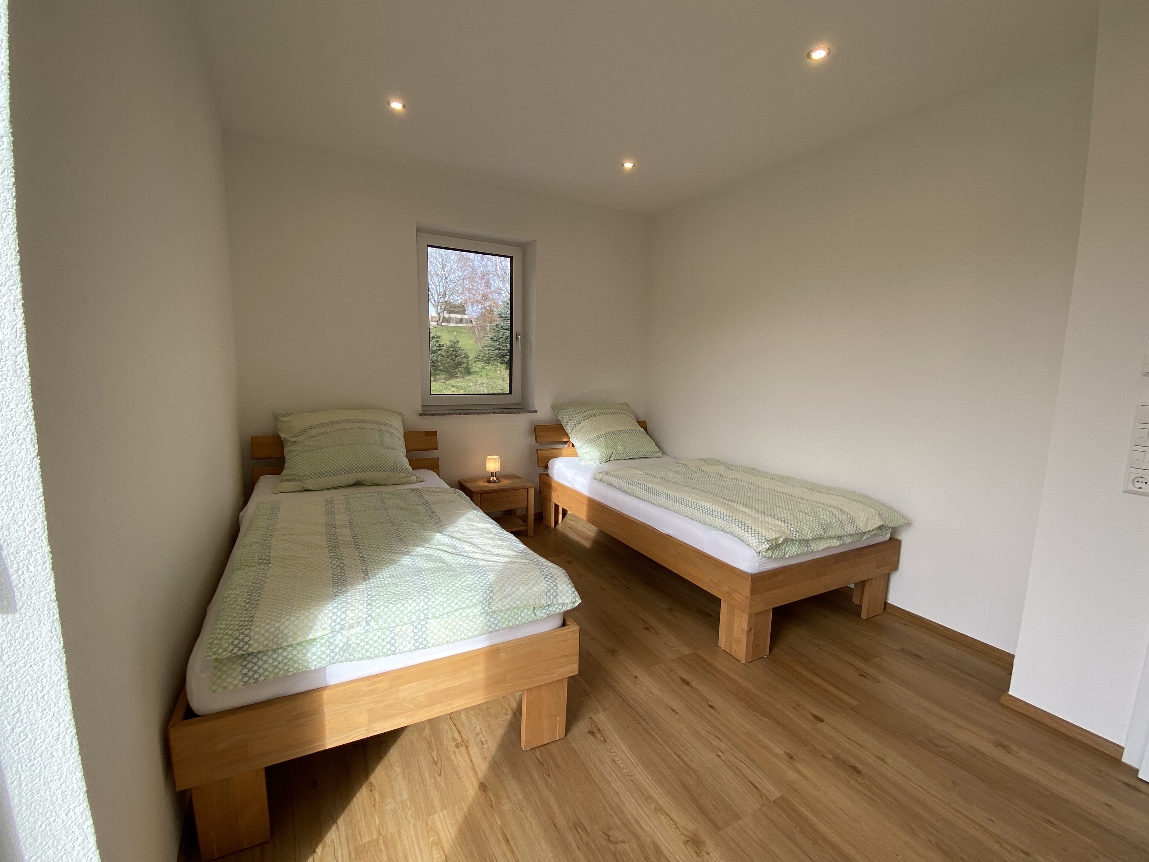 Een slaapkamer met twee houten bedden, een klein nachtkastje met een lamp en een raam met uitzicht op bomen.