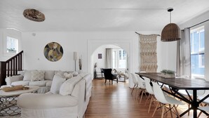 A spacious living room with a large sofa, dining table, and chairs. There is a staircase leading to another level, and a large window providing natural light. - Charming Home and Cottage in Downtown St Augustine for 14 (St. Augustine)