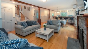 Ein geräumiges Wohnzimmer mit zwei grauen Sofas, einem weißen Couchtisch und einem Essbereich mit Tisch und Stühlen. Im Hintergrund ist eine Treppe zu einer weiteren Ebene zu sehen.