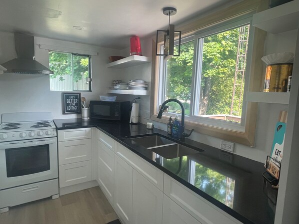 A modern kitchen with white cabinets, a black countertop, a stove, a microwave, a sink, and a window with a view of trees.