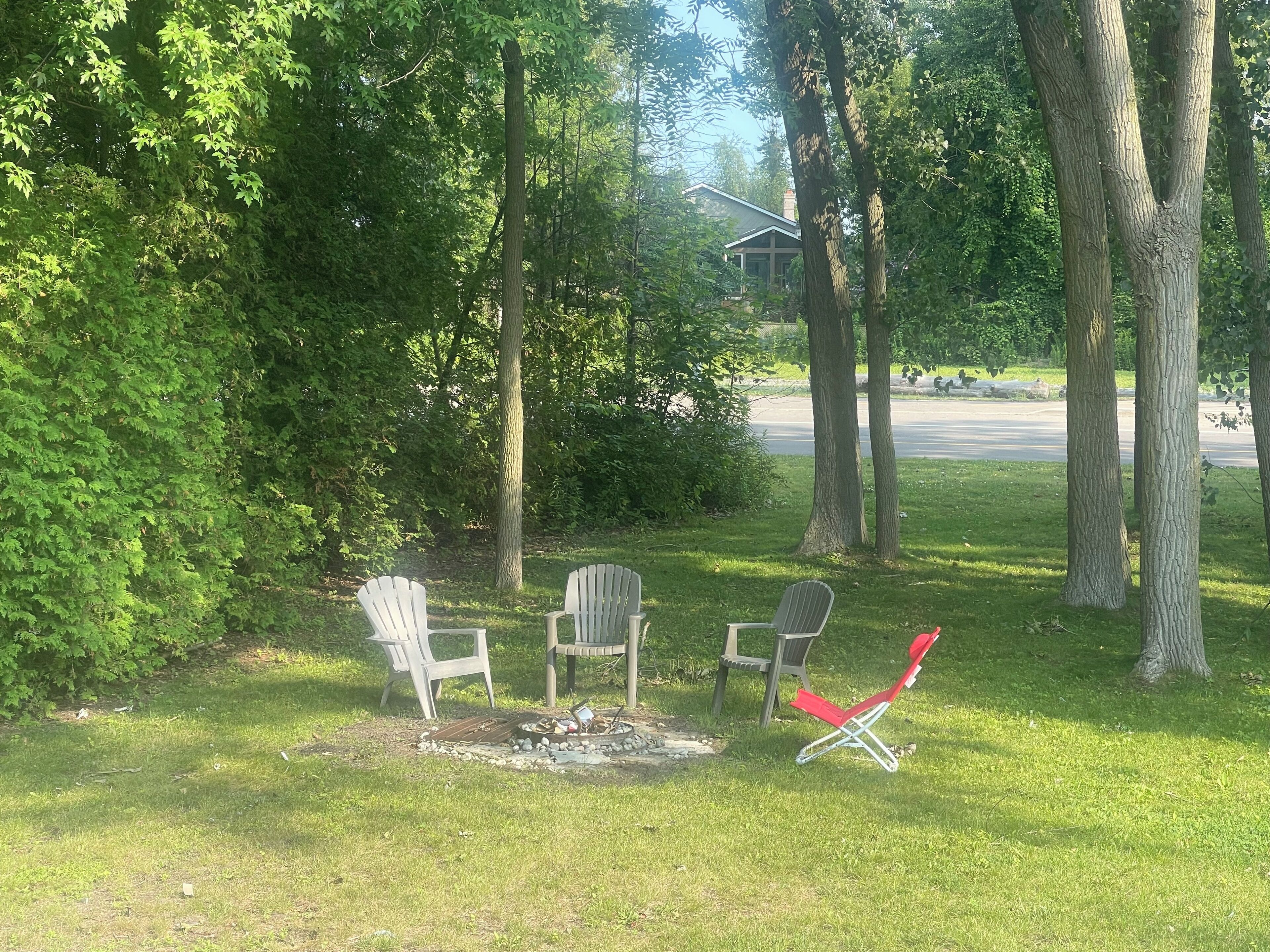 A grassy outdoor area with three Adirondack chairs, a fire pit, and a house in the background.