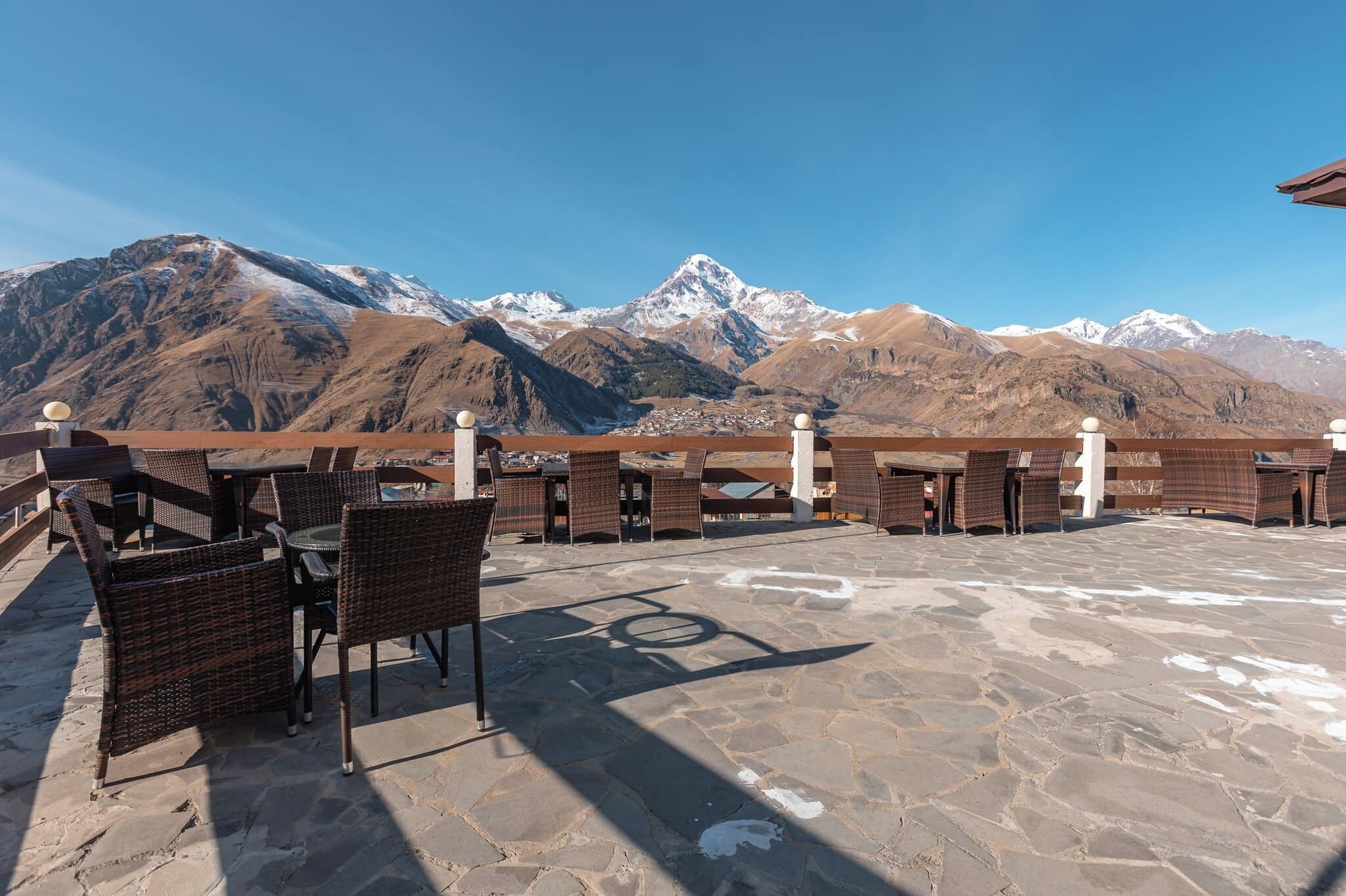 Een terras buiten met rieten meubilair en een rolstoeltoegankelijk pad, met uitzicht op besneeuwde bergen.