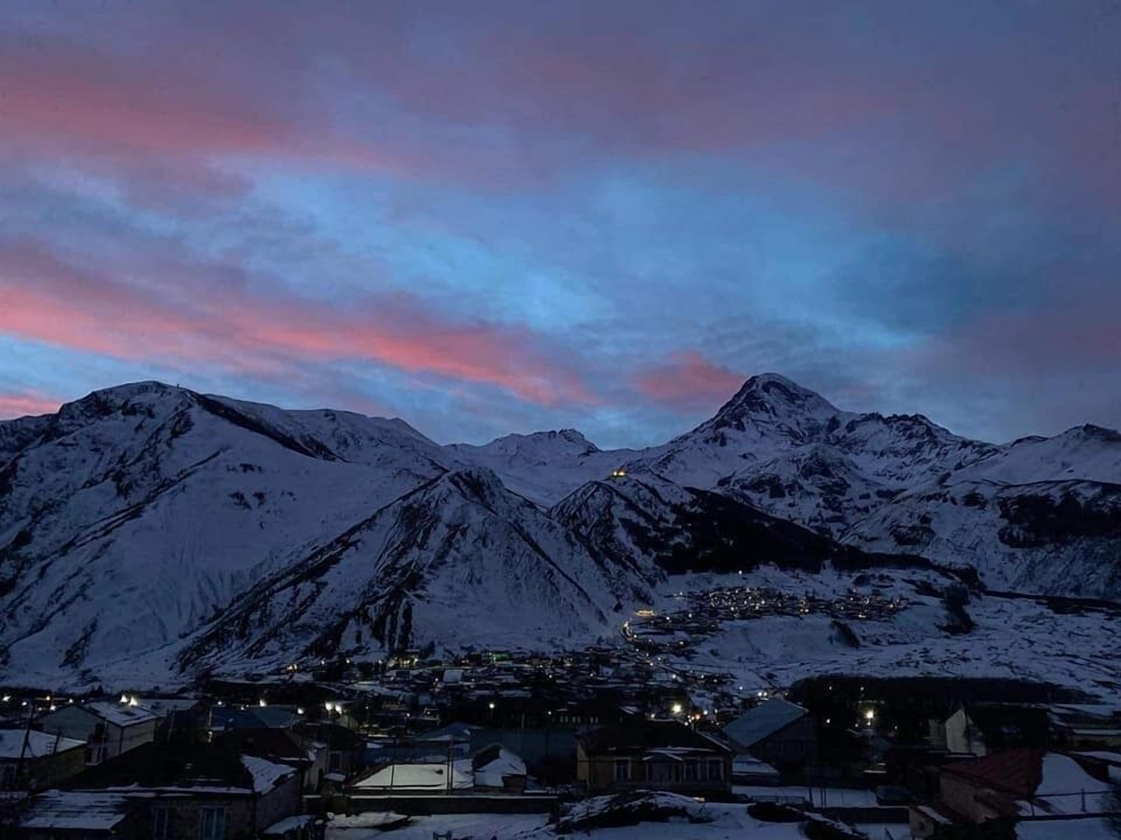 A snow-covered mountain range at dusk with a small settlement in the valley.