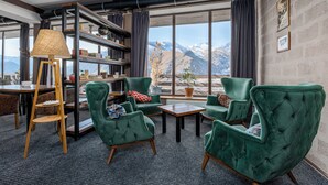 Ein gemütlicher Raum mit grün gepolsterten Sesseln, einem Holztisch und einem Bücherregal. Ein großes Fenster bietet Blick auf Berge.