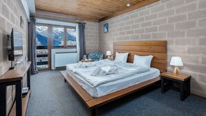 Ein Schlafzimmer mit Holzbalkendecke, einem großen Bett mit Kissen und Decke, einem Nachttisch mit Lampe, einem Fernseher auf einem Schrank und einem Fenster mit Bergblick.