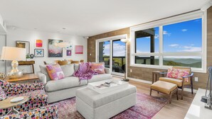 A modern living room with a grey sofa, a patterned ottoman, and a coffee table. There are several cushions on the sofa, and a large window offers a view of the outdoors. - Luxury Condo in the Sky! Finest in Wintergreen! (Wintergreen Resort)