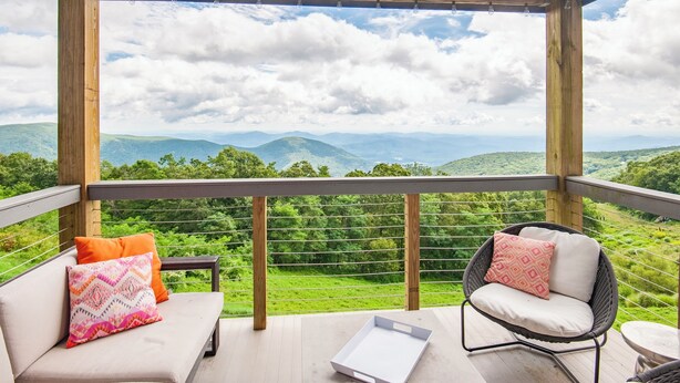 A wooden deck with a sofa and a wicker chair, overlooking a lush green landscape with mountains. - Luxury Condo in the Sky! Finest in Wintergreen! (Wintergreen Resort)
