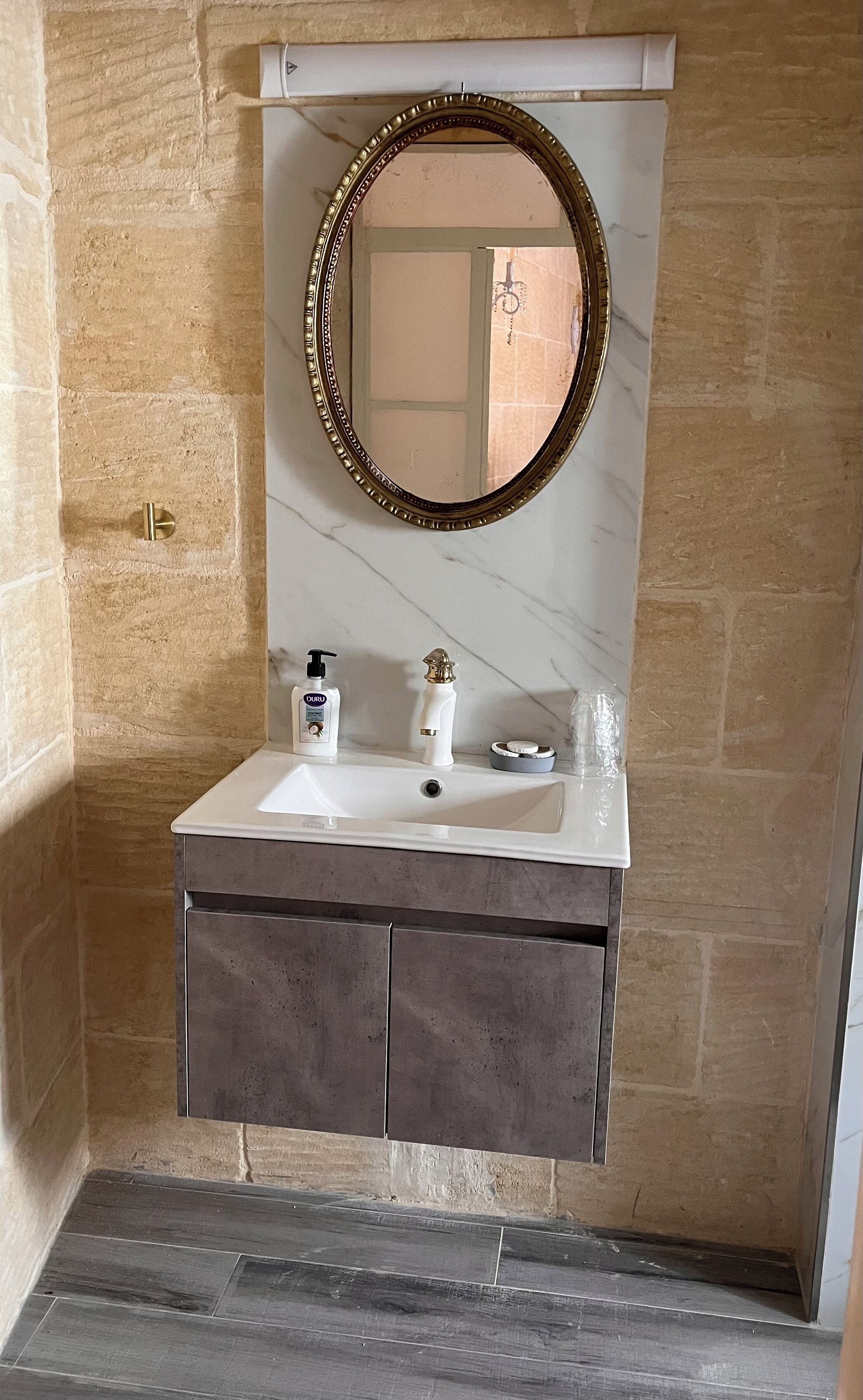 Un baño con lavamanos de mármol, espejo con marco dorado y pared de piedra.