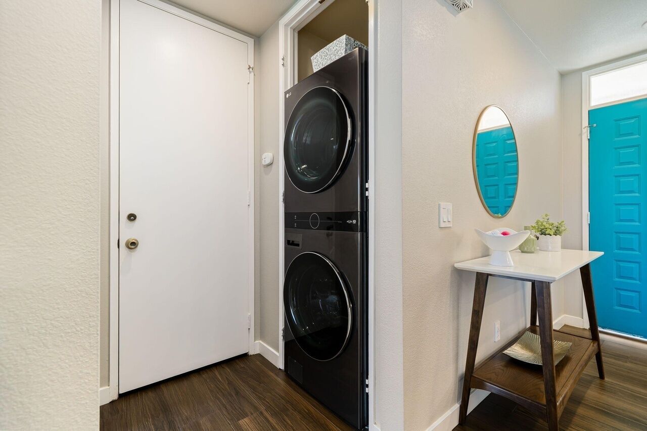 A laundry room with two stacked front-loading washing machines, a white door, a blue door, a small white table with a mirror, and a potted plant.