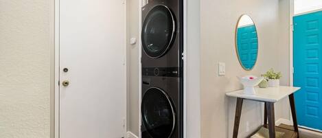 A laundry room with two stacked front-loading washing machines, a white door, a blue door, a small white table with a mirror, and a potted plant.