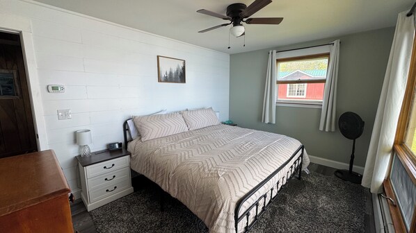 Un dormitorio con cama, mesita de noche, cómoda, ventilador de techo, ventana con cortinas y termostato en la pared.