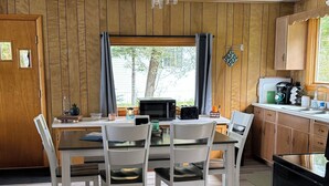 A dining area with a wooden table and chairs, a kitchen with a microwave and coffee maker, and a window with curtains.