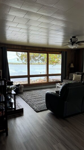 A living room with a large window overlooking a body of water, a ceiling fan, and a dark-colored sofa. - Relax in Aroostook County at this little piece of heaven right on the lake! (Portage Lake)
