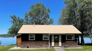 A small cabin with a metal roof, wooden siding, and a front porch. It is situated by a lake with trees in the background.
