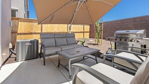 Une terrasse extérieure avec un parasol beige, un canapé gris, un barbecue et une table.