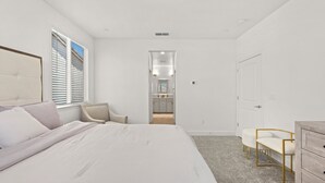 Une chambre avec un grand lit, une chaise, une commode et une vue sur la cuisine et la salle de bain, grâce à une porte ouverte.
