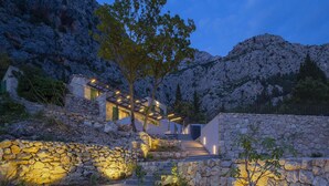 A modern villa with a stone facade, a staircase leading to the entrance, and a view of the surrounding rocky mountains.