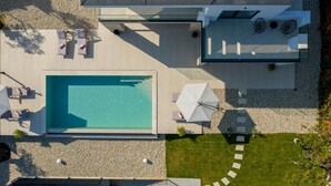 Luchtbeeld van een modern buitenzwembad met een rechthoekig zwembad, ligbedden, parasols en een pad dat leidt naar een overdekt zitgedeelte.