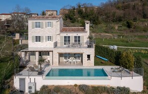 A two-story stone house with a swimming pool, outdoor seating, and a view of the surrounding landscape.
