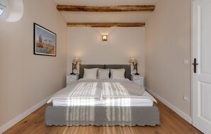 A bedroom with a bed, two bedside tables, a nightstand, a wall-mounted lamp, a door, and a framed picture on the wall.