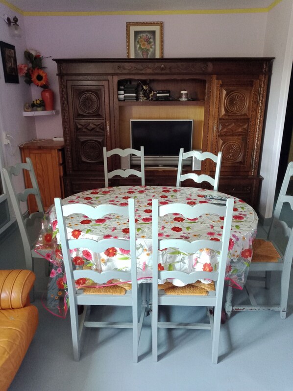 A dining area with a round table covered in a floral tablecloth, surrounded by white chairs with woven seats. - Maison Complète (Montluçon)