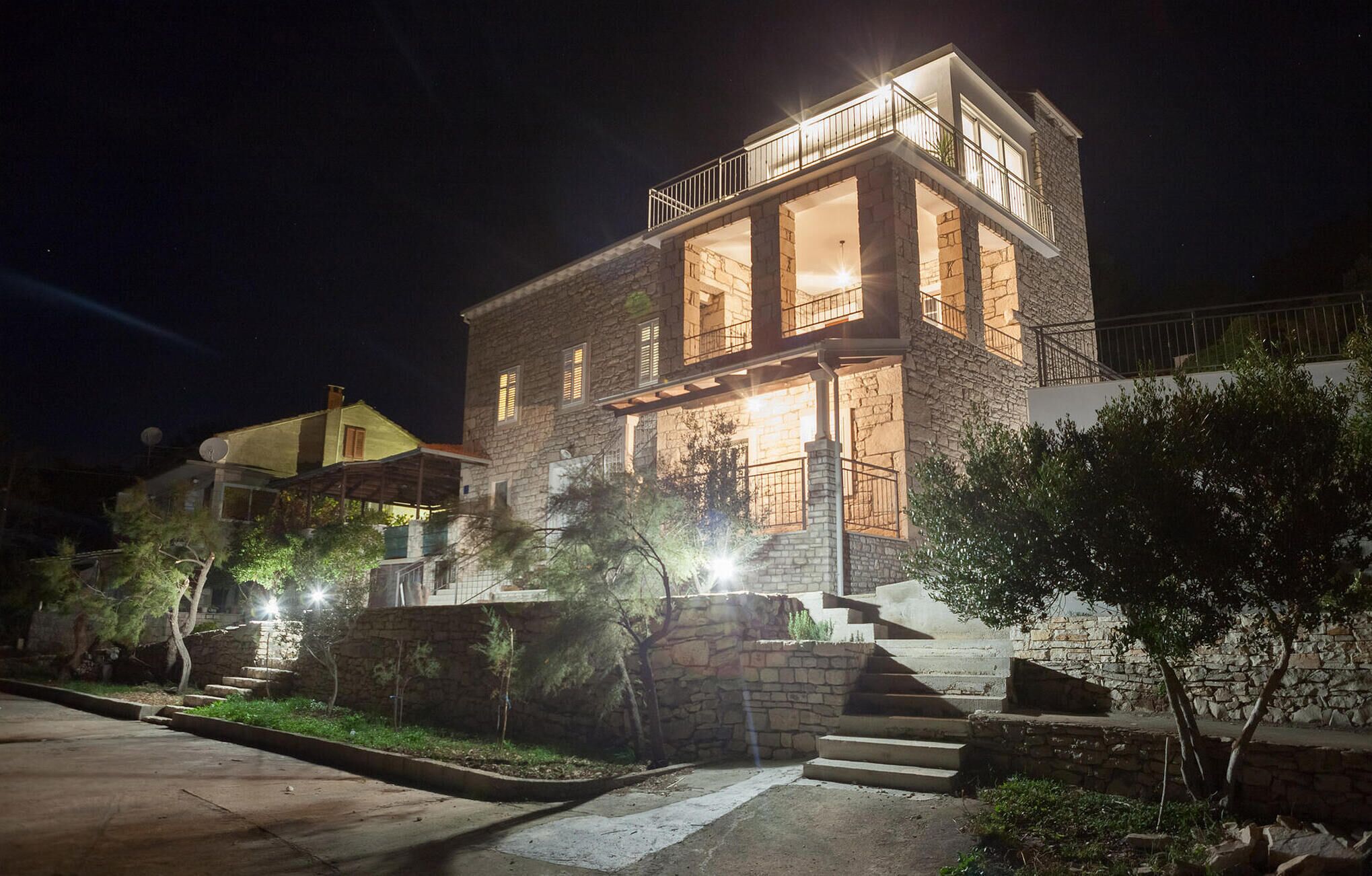 A two-story stone house with a balcony, illuminated windows, and a staircase leading to the entrance.