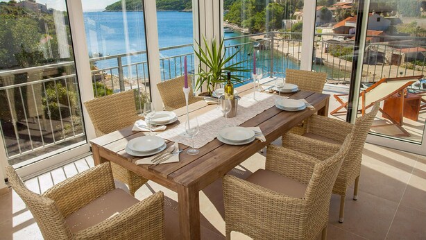 A dining area with a wooden table set for six, surrounded by wicker chairs, overlooking a scenic coastal view.