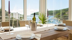 A dining table set with plates, cutlery, and a bottle of wine, overlooking a scenic view of the sea and hills.