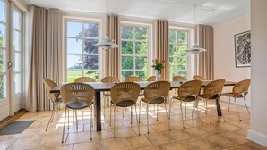 Une salle à manger avec une longue table, de nombreuses chaises, de grandes fenêtres avec des rideaux et un vase rempli de fleurs.