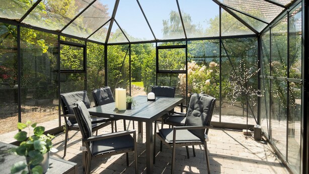 Une serre avec un toit en verre, une table avec des chaises et des plantes en pot.