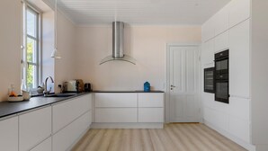 Une cuisine moderne avec des meubles blancs, une hotte en acier inoxydable, un four encastré et une fenêtre qui laisse entrer la lumière naturelle.