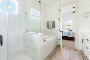 A modern bathroom with a glass-enclosed shower, a freestanding bathtub, and a visible bedroom with a bed and a window.
