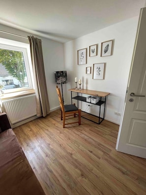 A room with a wooden floor, a desk with a chair, a radiator, a window with curtains, and framed pictures on the wall.