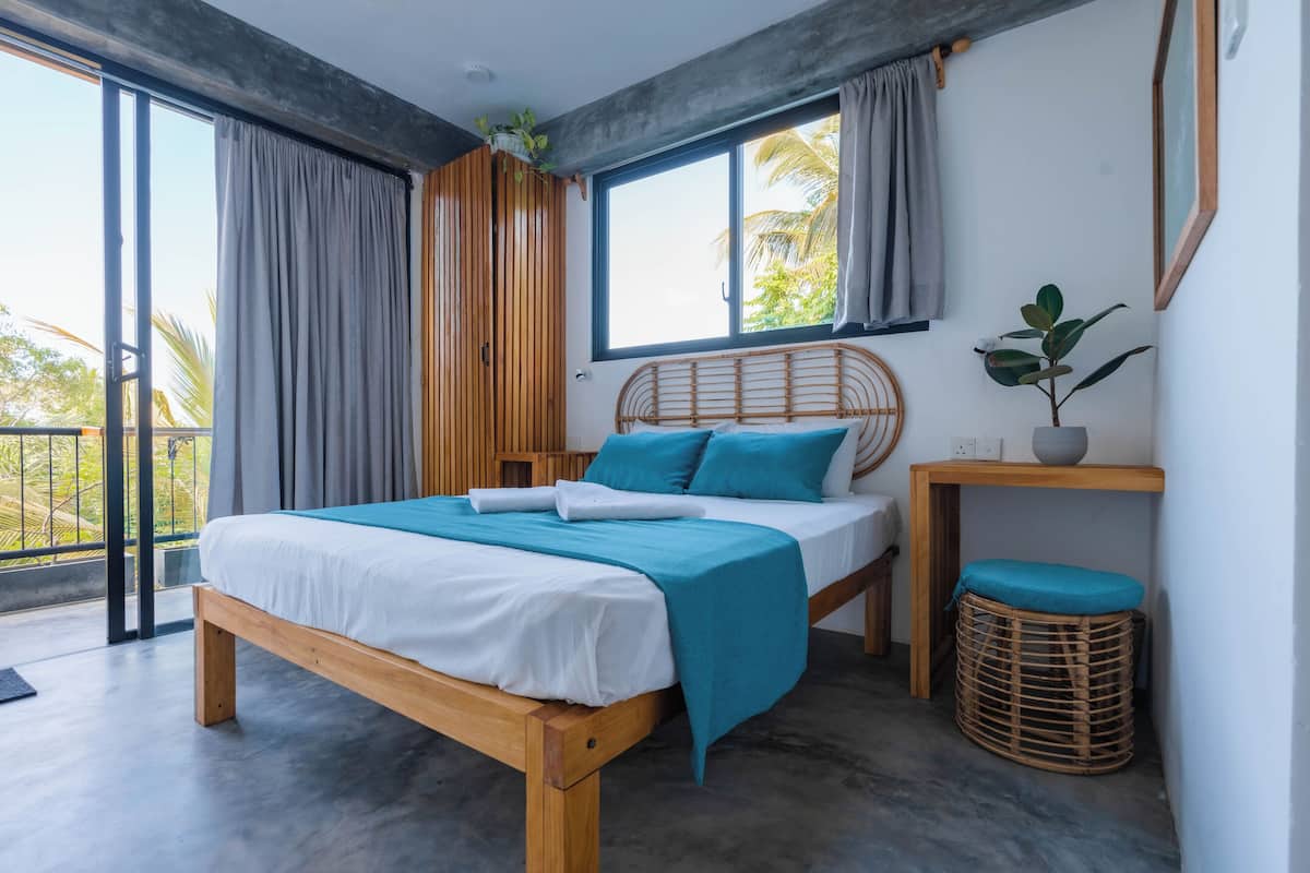 A modern bedroom with a wooden bed, a small table with a plant, and a view of palm trees through large windows.