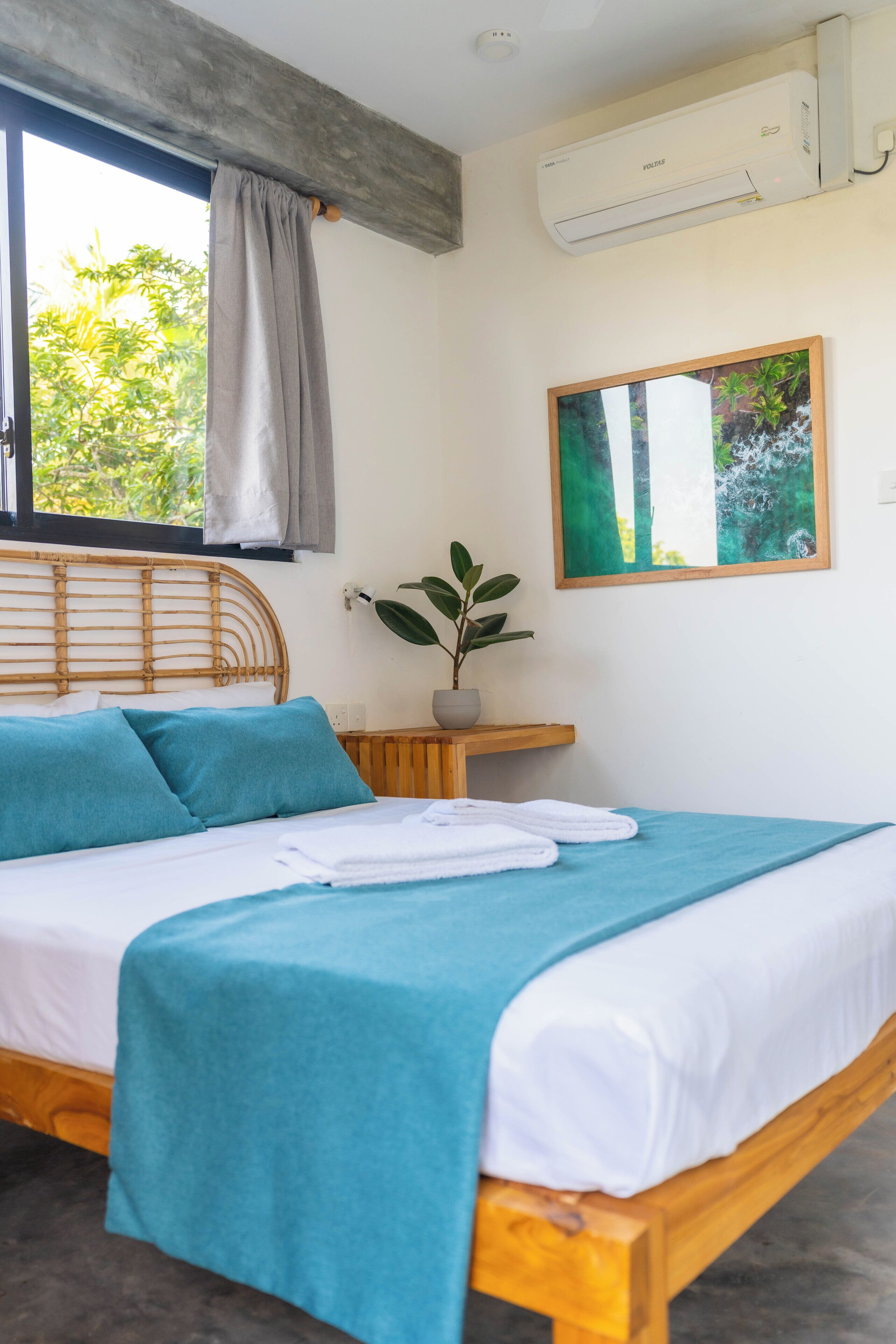 A bed with a wooden frame, a blue bedspread, and white bedding. There is a window with a view of greenery outside.