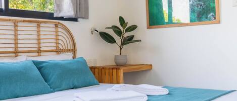 A bed with a wooden frame, a blue bedspread, and white bedding. There is a window with a view of greenery outside.