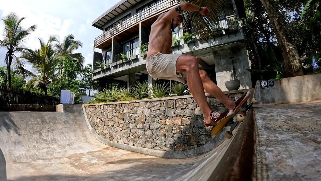Un homme effectue une figure de skateboard sur une rampe en béton, avec un mur de pierres et un bâtiment en arrière-plan.