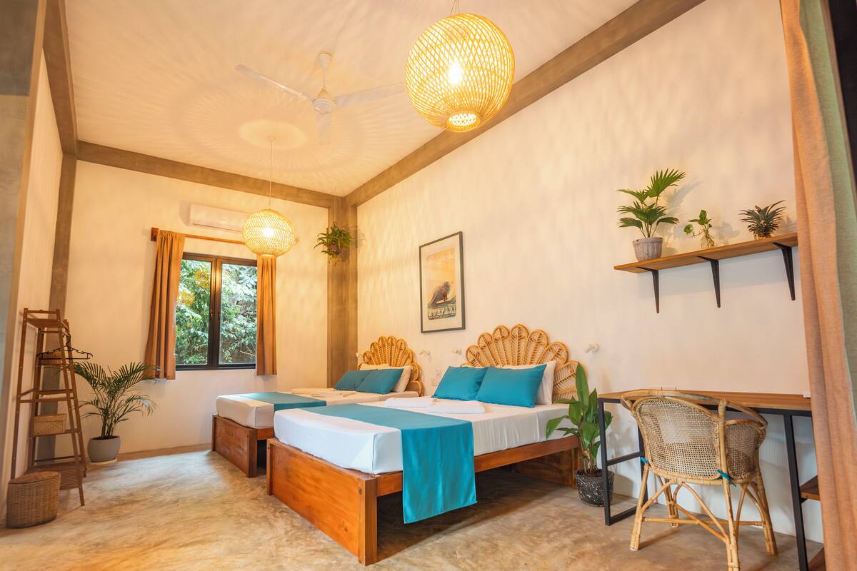 A bedroom with two beds, a desk, wicker furniture, and potted plants.
