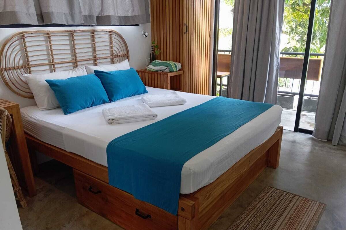 A bed with a wooden frame, white bedding, and blue pillows. There is a wooden bench and a small table with a plant beside the bed.