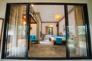 A spacious room with a large sliding glass door leading to an outdoor area. The room has a bed with blue bedding, a sofa, and a small table. There is a ceiling fan and a hanging light fixture.