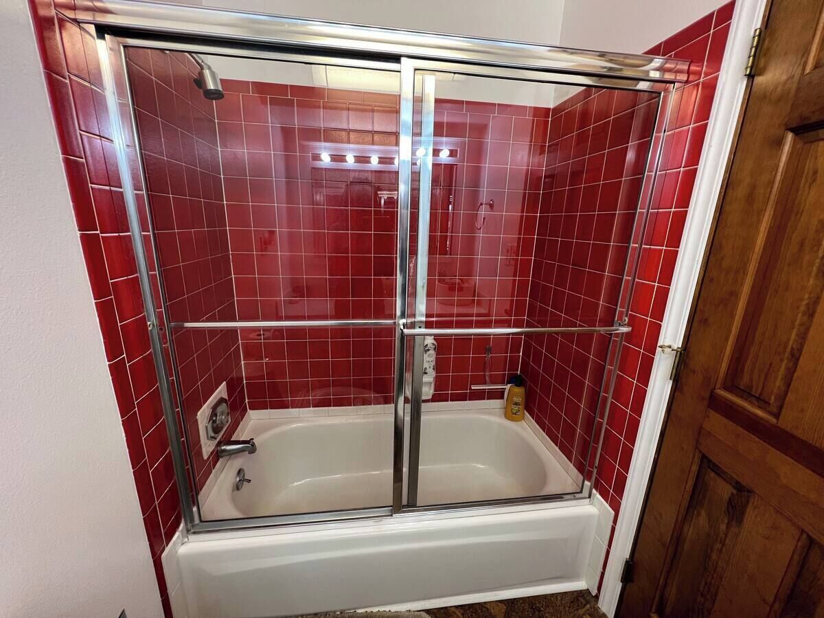 Eine begehbare, gläserne Dusche mit einer weißen Badewanne, roten Fliesenwänden und einer Holztür.