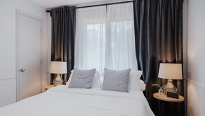 Een net opgemaakt bed met witte lakens, twee bedlampjes en een klein tafeltje met een vaas en een boek.