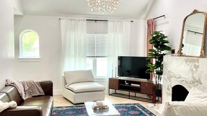 A modern living room with a brown leather sofa, a white armchair, a TV, a marble fireplace, and a chandelier.