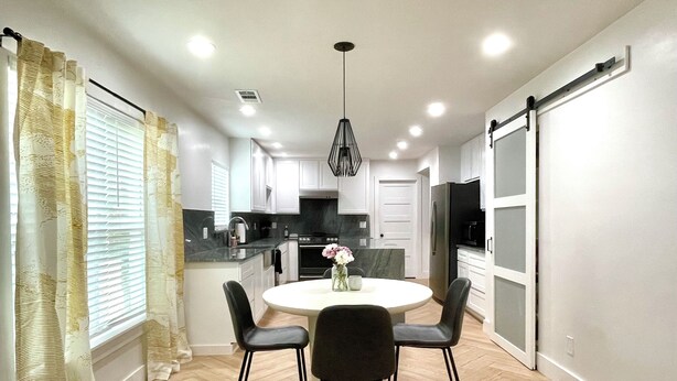 A modern kitchen with a dining area, featuring a round table with chairs, a hanging light fixture, and a sliding door.