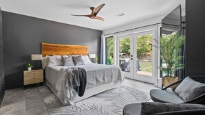 Un dormitorio moderno con una cama grande, un ventilador de techo, una mesita de noche con lámpara, un sofá gris y vistas a un jardín a través de puertas de cristal.